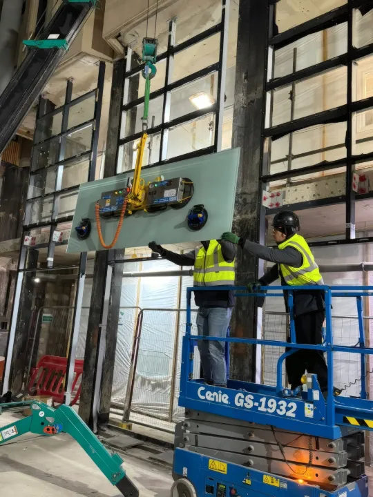 A glazing unit lifted into the facade frame on a vacuum lifter, guided by two installers.