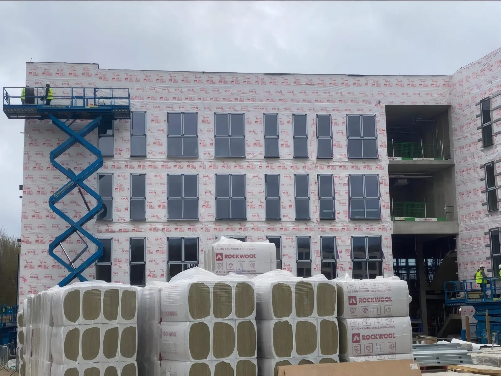 A scissor lift at the membrane-wrapped elevation, with insulation deliveries in front.